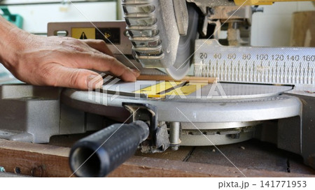 A craftsman works at a miter saw in his workshop. His hand secures a thin block of wood to the tool frame before cutting. The equipment features a clearly visible scale for precise and safe wood cut A craftsman works at a miter saw in his workshop. His hand secures a thin block of wood to the tool frame before cutting. The equipment features a clearly visible scale for precise and safe wood cut 141771953