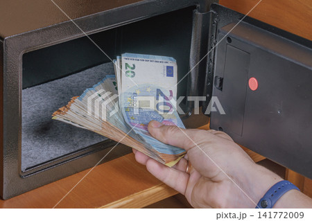 Person hand taking euro cash banknotes out of open safe box placed on wooden shelf inside hotel room cabinet for financial security asset storage concept Person hand taking euro cash banknotes out of open safe box placed on wooden shelf inside hotel room cabinet for financial security asset storage concept 141772009