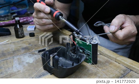 A mechanic repairs a battery pack in a workshop. He holds a soldering iron, carefully soldering contacts to the electronic circuit board with wires, and a gray cable, working at a workbench. 141772020