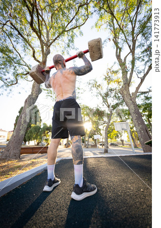 Man lifts heavy weights in a park under the morning sun with trees surrounding the area Man lifts heavy weights in a park under the morning sun with trees surrounding the area 141773913
