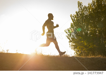 Runner moves along a path in the sunlight during late afternoon near trees Runner moves along a path in the sunlight during late afternoon near trees 141773947