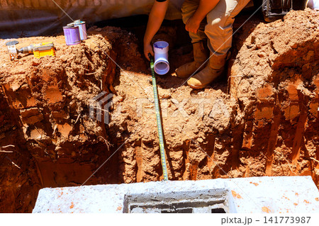 Worker is placing pipe into trench at construction site while measuring it with tape measure Worker is placing pipe into trench at construction site while measuring it with tape measure 141773987