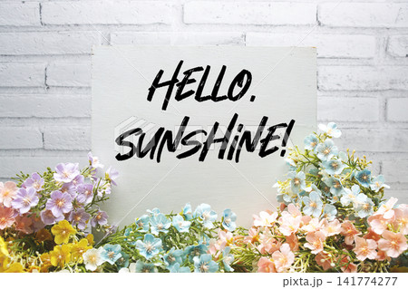"Hello, Sunshine!" Message surrounded by colorful pastel flowers on a wooden surface, set against a white brick wall 141774277