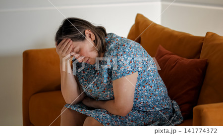 Woman Experiencing Pain While Seated Silently. Woman Shows Signs Of Stress And Headache Indoors Quietly. Woman Sitting On Sofa Exhibits Tension And Discomfort Due To Headache In Peaceful Room Woman Experiencing Pain While Seated Silently. Woman Shows Signs Of Stress And Headache Indoors Quietly. Woman Sitting On Sofa Exhibits Tension And Discomfort Due To Headache In Peaceful Room 141774372