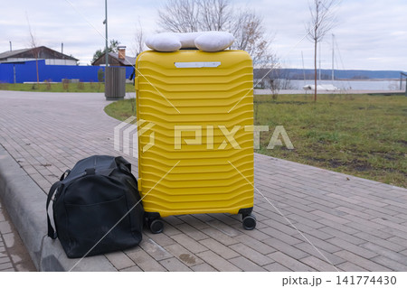 Yellow Suitcase Crowned By Travel Pillow Near Path With Duffel Beside, Restful Park Stop With Grassy Bank And Distant Water, Casual Resting Moment For Traveler Between Legs Of Journey Yellow Suitcase Crowned By Travel Pillow Near Path With Duffel Beside, Restful Park Stop With Grassy Bank And Distant Water, Casual Resting Moment For Traveler Between Legs Of Journey 141774430