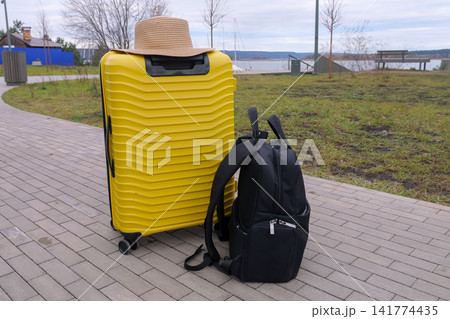 Yellow Suitcase With Straw Hat On Path Beside Waterfront, Black Backpack Leaning Against Trolley, Overcast Sky And Grassy Bank Set Anticipatory Mood, Readyforescape Scene For Weekend Getaway Yellow Suitcase With Straw Hat On Path Beside Waterfront, Black Backpack Leaning Against Trolley, Overcast Sky And Grassy Bank Set Anticipatory Mood, Readyforescape Scene For Weekend Getaway 141774435