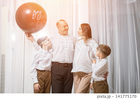 Caucasian Family Celebrating Gender Party Boy, Father Lifting Balloon Overhead, Mother Looking At Father, Child Reaching Up, Warm Backlight Through Drapes, Lively Candid Action, Cheerful Announcement 141775170