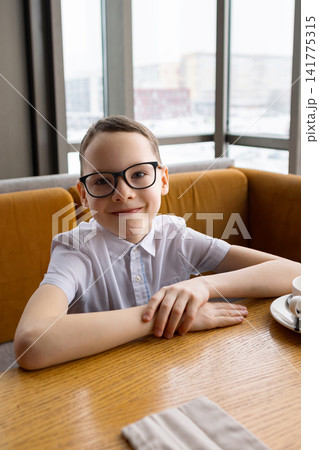 Young Boy Sitting At Table Near Window Wearing Glasses And White Shirt, Smiling At Camera Over Cup And Plate, Warm Natural Light Filling Modern Cafe Interior, Candid Student Portrait Conveying Young Boy Sitting At Table Near Window Wearing Glasses And White Shirt, Smiling At Camera Over Cup And Plate, Warm Natural Light Filling Modern Cafe Interior, Candid Student Portrait Conveying 141775315