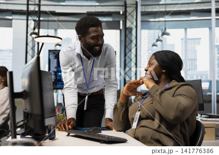 Jolly programmers in office enjoying conversation while reviewing code snippets, adjusting configurations. Smiling african american coworkers chatting while running diagnostic tools on PC 141776386