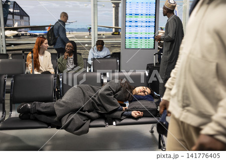 Jet lagged woman sleeping on airport bench while waiting for flight, passing time. Bored girl taking nap on departure hub chairs while waiting for plane taking her to travel destination Jet lagged woman sleeping on airport bench while waiting for flight, passing time. Bored girl taking nap on departure hub chairs while waiting for plane taking her to travel destination 141776405