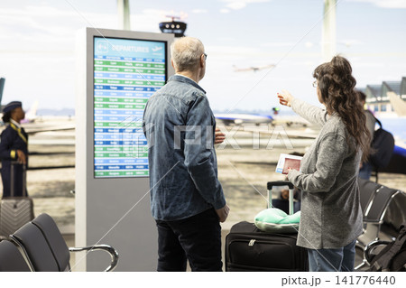 Happy senior couple looking at departure information on large digital screen, standing together in airport terminal. Elderly travelers checking flight details and boarding status in airport. Happy senior couple looking at departure information on large digital screen, standing together in airport terminal. Elderly travelers checking flight details and boarding status in airport. 141776440