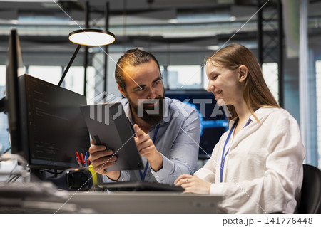 Cheerful technician mentoring young employee in startup office, reviewing code scripts on tablet. Jolly tech company team leader guiding intern doing software development Cheerful technician mentoring young employee in startup office, reviewing code scripts on tablet. Jolly tech company team leader guiding intern doing software development 141776448