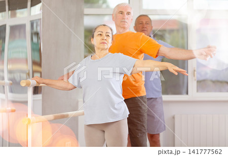 Dancing positive elderly women and men engaged in group class perform exercise near ballet barre, standing in 2nd position of ballet stance in dance studio 141777562