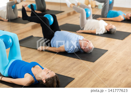 People of different ages pressing core and clutching mini Pilates balls between legs during Pilates training together People of different ages pressing core and clutching mini Pilates balls between legs during Pilates training together 141777623