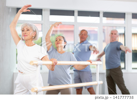 Experienced mature female ballet dancer, dressed in sportswear and group of women and men engaged in classical ballet performs exercise standing in 3rd position near ballet barre in dance hall Experienced mature female ballet dancer, dressed in sportswear and group of women and men engaged in classical ballet performs exercise standing in 3rd position near ballet barre in dance hall 141777738