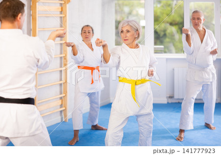 Group of elderly people in white kimonos learn to punch and kick under the guidance of trainer. Karate lessons Group of elderly people in white kimonos learn to punch and kick under the guidance of trainer. Karate lessons 141777923