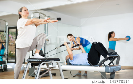Aged woman practicing pilates on pedal fitness chair in sports hall Aged woman practicing pilates on pedal fitness chair in sports hall 141778114