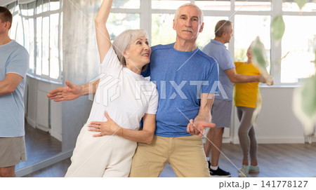 Positive senior woman and man practicing tango dance moves as a couple during a group celebration in dance studio 141778127