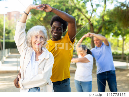 Exited older and middle-aged diverse people enjoying latin dances breaking into pairs on sandy area between trees Exited older and middle-aged diverse people enjoying latin dances breaking into pairs on sandy area between trees 141778135