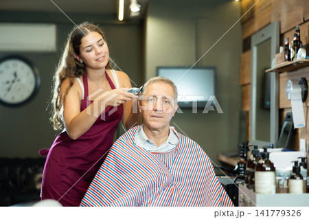 Young woman hairdresser performs haircut for elderly man using hair clipper Young woman hairdresser performs haircut for elderly man using hair clipper 141779326