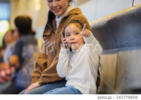 child girl talking on a smartphone and pointing with something with her mother while sitting on bench at a bus station in city at night child girl talking on a smartphone and pointing with something with her mother while sitting on bench at a bus station in city at night 141779859