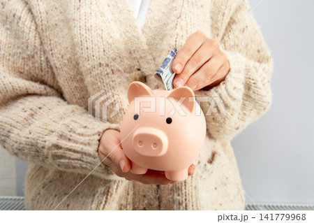 Heating season. Person inserts cash into piggy bank wearing cozy sweater Heating season. Person inserts cash into piggy bank wearing cozy sweater 141779968