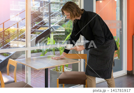 Young male waiter or barista in striped apron wiping and cleaning wooden table while holding coffee cup in modern cafe restaurant Young male waiter or barista in striped apron wiping and cleaning wooden table while holding coffee cup in modern cafe restaurant 141781917