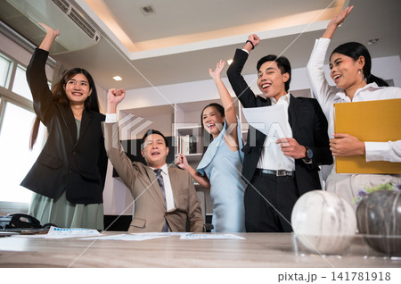 Excited business team celebrating success in modern office and raising hands with joyful. Asian professionals in formal wears expressing triumph after achieving goals or closing deal. 141781918