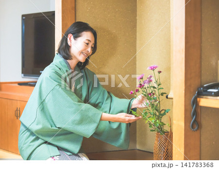 温泉旅館の床の間で生け花に手を添え笑みを浮かべる浴衣姿の女性 温泉旅館の床の間で生け花に手を添え笑みを浮かべる浴衣姿の女性 141783368
