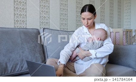 Young mother multitasking working on laptop holding baby Young mother multitasking working on laptop holding baby 141784164