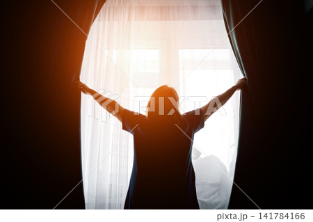 Woman opening curtains in front of a bright window with soft backlight Woman opening curtains in front of a bright window with soft backlight 141784166