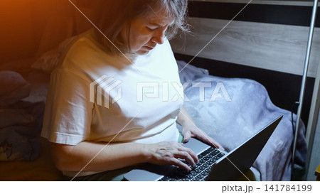 Mature woman in pajamas typing on a laptop in bed at night, bathed in screen glow, working or browsing online late at home, comfortable and connected in bedroom light Mature woman in pajamas typing on a laptop in bed at night, bathed in screen glow, working or browsing online late at home, comfortable and connected in bedroom light 141784199