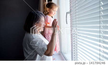 Grandmother and little girl standing together at a window with blinds, waving goodbye and showing feelings of separation, connection, social distancing, and family love Grandmother and little girl standing together at a window with blinds, waving goodbye and showing feelings of separation, connection, social distancing, and family love 141784246