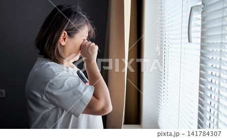 Mature woman standing by a window with closed blinds, hand covering her face as she cries, conveying solitude, grief and quiet emotional pain in a dim domestic interior Mature woman standing by a window with closed blinds, hand covering her face as she cries, conveying solitude, grief and quiet emotional pain in a dim domestic interior 141784307