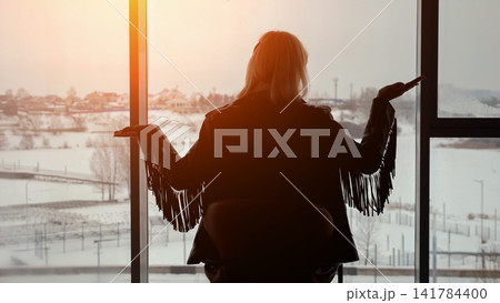Woman silhouette looking out of a large window with open arms, observing the serene winter landscape and orange light from the setting sun, feeling freedom and contemplation Woman silhouette looking out of a large window with open arms, observing the serene winter landscape and orange light from the setting sun, feeling freedom and contemplation 141784400