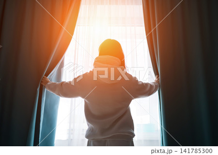 Person standing with back to camera, opening curtains at a window, letting bright morning light flood into the room, symbolizing a fresh start and hope Person standing with back to camera, opening curtains at a window, letting bright morning light flood into the room, symbolizing a fresh start and hope 141785300