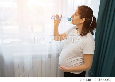 Pregnant woman standing on a digital scale in a bright room with dumbbells, resistance band, and water bottle nearby, wellness and home fitness scene Pregnant woman standing on a digital scale in a bright room with dumbbells, resistance band, and water bottle nearby, wellness and home fitness scene 141785301