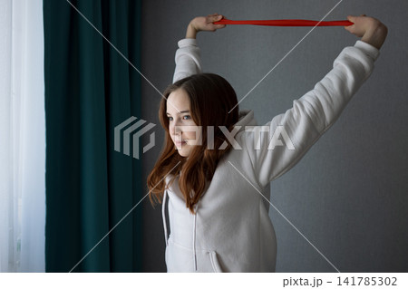 Young woman in a white hoodie performing resistance band exercises, stretching her arms above her head, focusing on fitness and wellness in a comfortable home environment 141785302