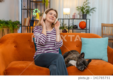 Middle-aged woman with dog wears wireless headphones listens to music and dances rhythm energy happy Middle-aged woman with dog wears wireless headphones listens to music and dances rhythm energy happy 141785731