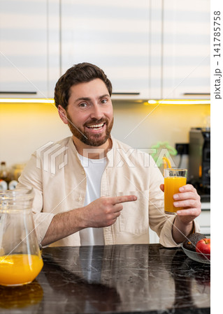 Middle-aged man smiling pointing at orange juice glass in home kitchen promoting healthy diet drink Middle-aged man smiling pointing at orange juice glass in home kitchen promoting healthy diet drink 141785758