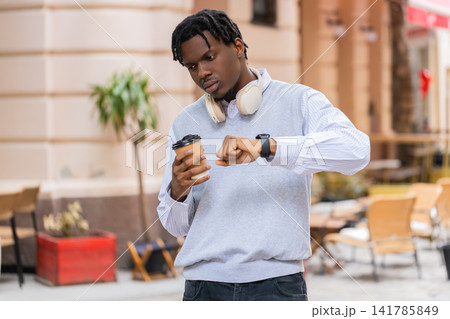 African American young man holding coffee walking in city street checking watch and feeling annoyed African American young man holding coffee walking in city street checking watch and feeling annoyed 141785849