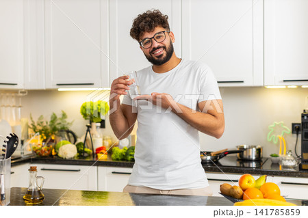 Arabian young adult man drinking water in kitchen at home promoting hydration habit for fit wellness 141785859