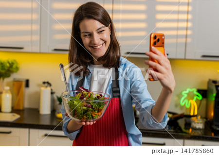 Young woman filming healthy salad vlog on smartphone in home kitchen for diet nutrition lifestyle Young woman filming healthy salad vlog on smartphone in home kitchen for diet nutrition lifestyle 141785860