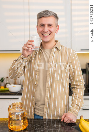 Middle-aged man drinking water in kitchen at home with smile after diet meal for weight loss routine Middle-aged man drinking water in kitchen at home with smile after diet meal for weight loss routine 141786251