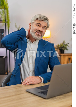 Middle-aged businessman grimaces sudden neck pain clutching neck seeking relief during work session Middle-aged businessman grimaces sudden neck pain clutching neck seeking relief during work session 141786287