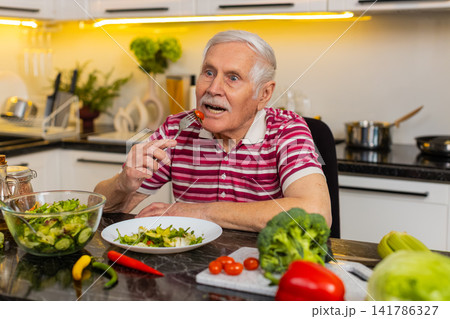 Senior old man happily eating fresh vegetable salad in home kitchen for healthy diet and weight loss 141786327