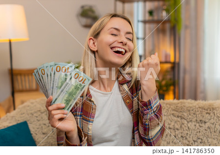 Adult woman waving stack of dollar cash at home smiling with joy celebrating major financial success 141786350