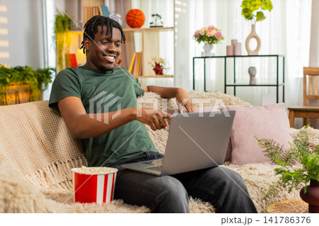 African American young man on home sofa watching laptop with popcorn feeling surprised and amused African American young man on home sofa watching laptop with popcorn feeling surprised and amused 141786376