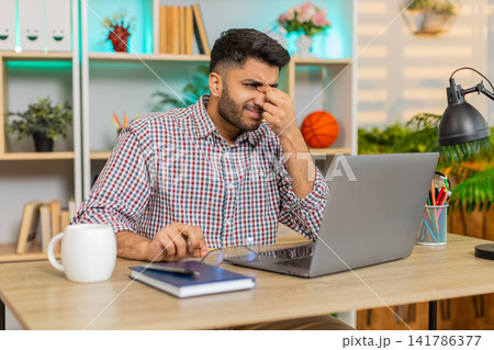 Indian man on home table rubbing tired eyes from laptop work and feeling fatigue during remote job 141786377