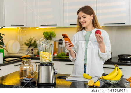 Middle-aged woman choosing fruit for smoothie using smartphone in home kitchen for healthy diet plan Middle-aged woman choosing fruit for smoothie using smartphone in home kitchen for healthy diet plan 141786454